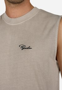 Ermangeløs beige skjorte uden ærmer lavet af blødt stof, med en ribbet hals og sort broderet "Pegador" logo på brystet. Simpelt design.