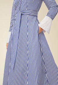 Abito a righe blu e bianche con maniche lunghe e polsini bianchi, una cintura coordinata e una vestibilità ampia con una gonna a ruota e tasche.