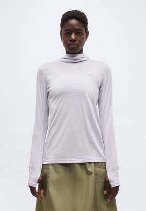 Άτομο που φοράει ανοιχτό μωβ μακρυμάνικο ζιβάγκο και φούστα ελιάς, στέκεται μπροστά σε απλό λευκό φόντο.