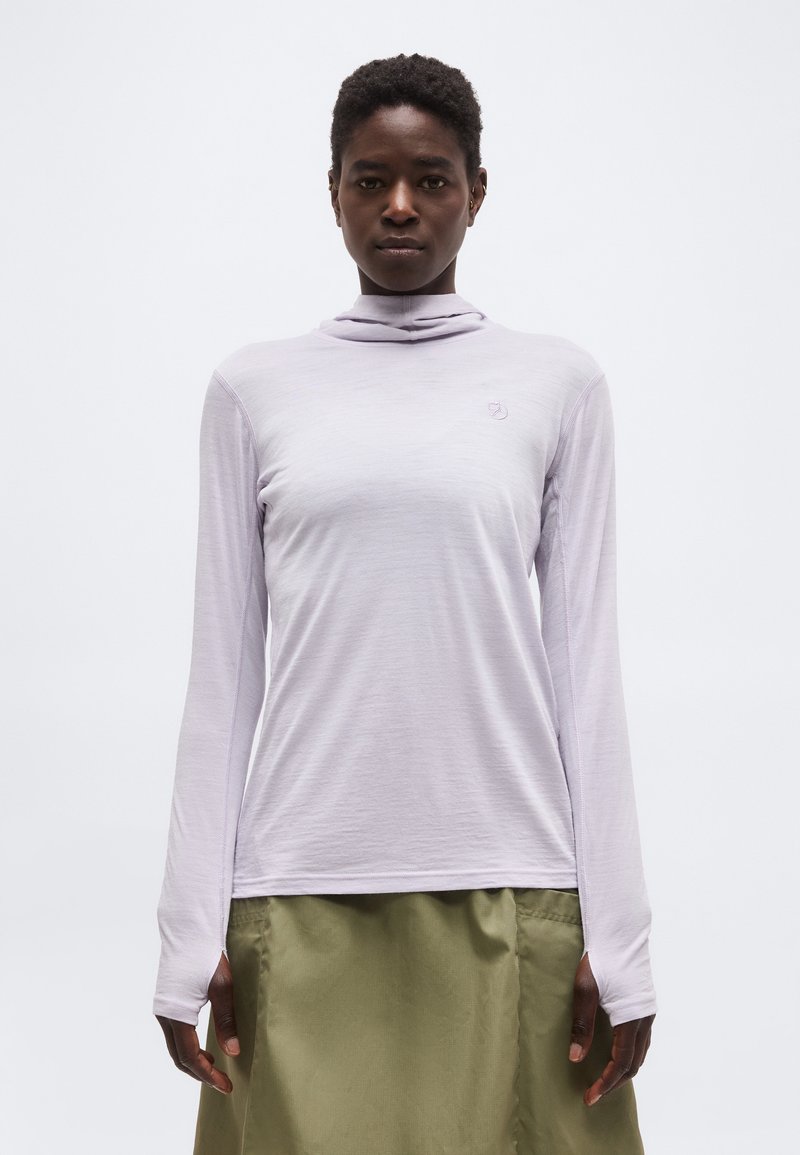 Personne portant un col roulé à manches longues couleur lavande claire et une jupe vert olive, debout devant un fond blanc uni.