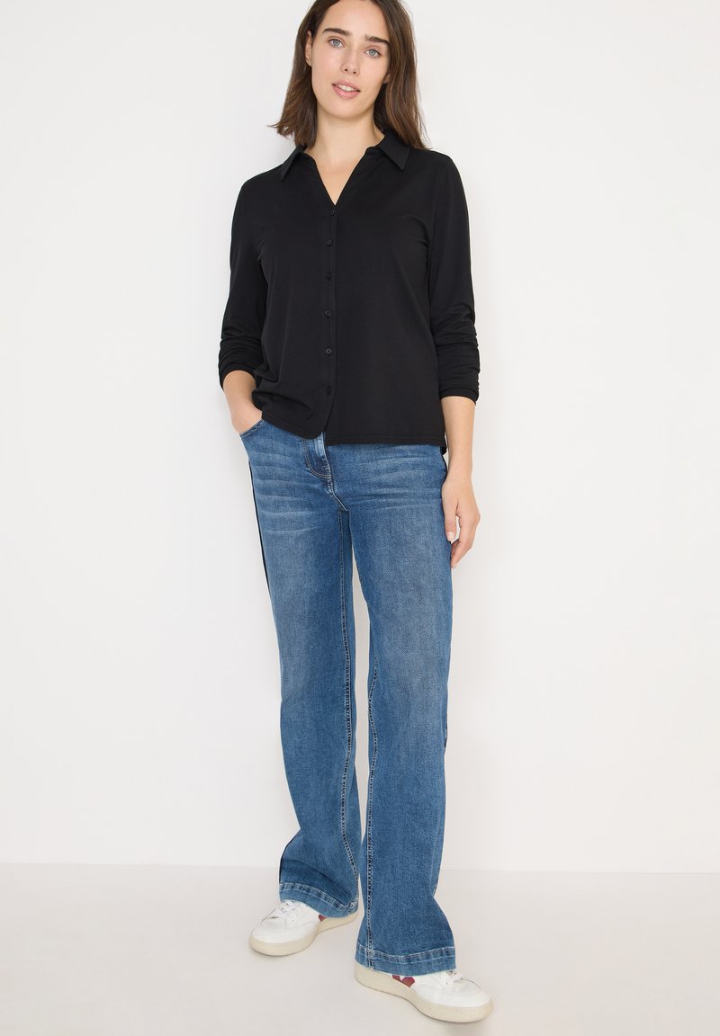 Camisa negra de botones con mangas largas, combinada con jeans azules de campana y zapatillas blancas. Diseño simple, ajuste casual.