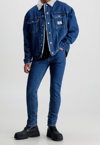 Jeansjacka med lammullskrage, blåtvätt, två bröstfickor; kombinerad med blå tighta jeans och kraftiga svarta kängor.