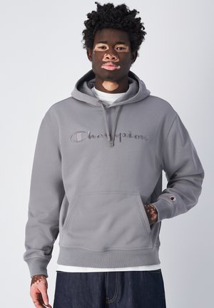 Sudadera gris con cordón y bolsillo frontal, con el logotipo "Champion" bordado. Puños y dobladillo acanalados. Worn sobre una camiseta blanca.