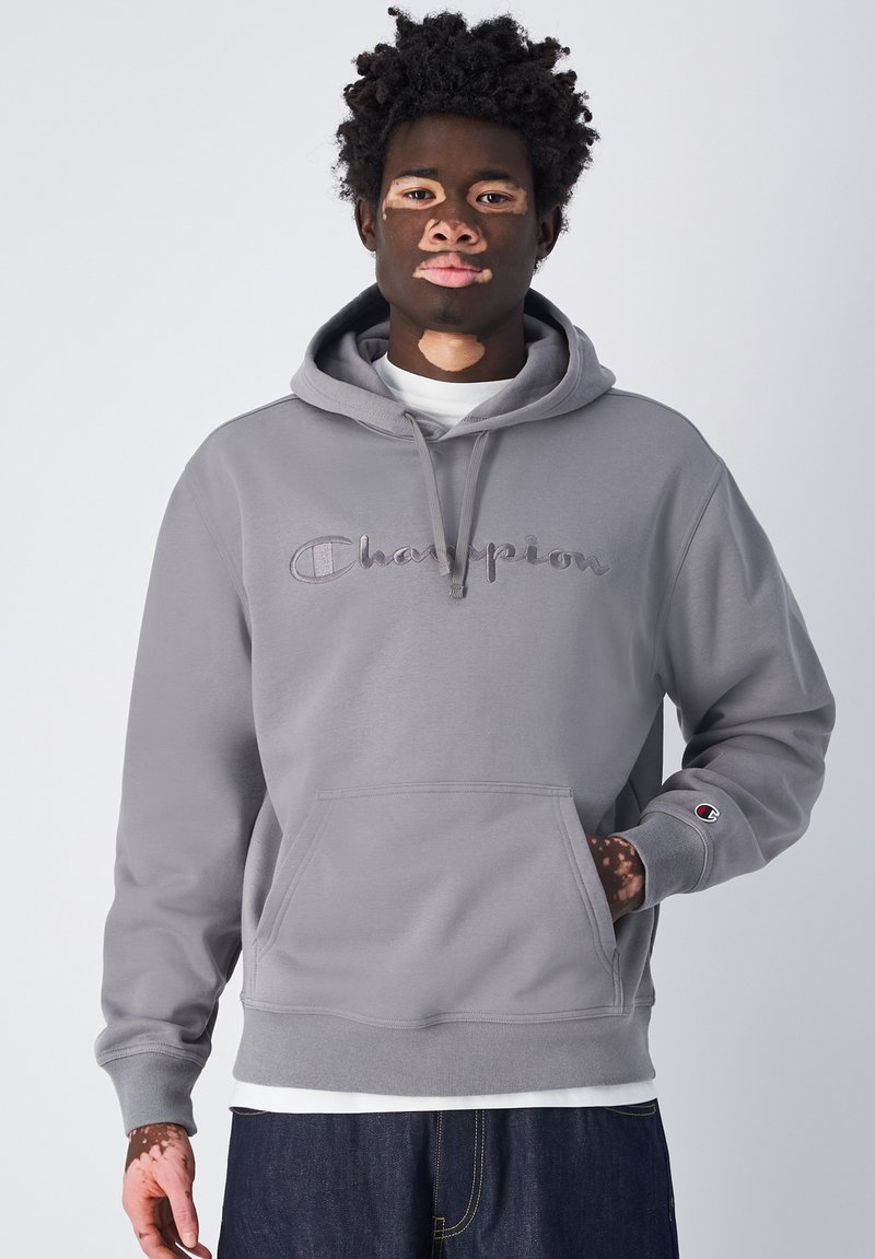 Grau Kapuzenpullover mit Kordelzug und Fronttasche, mit gesticktem "Champion"-Logo. Gerippte Bündchen und Saum. Über einem weißen Shirt getragen.