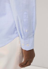 Puño de camisa azul claro con dos botones de perla, que presenta una tela suave y texturizada y un diseño de puño puntiagudo. Detalles de costura visibles.