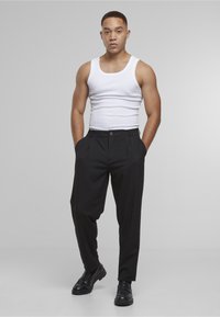 Svarte plisserte bukser med glatt tekstur, frontlommer og knappelukking. Kombinert med en tettsittende hvit singlet og svarte sko.