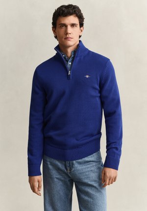 Homme portant un pull bleu à fermeture éclair jusqu'à la poitrine par-dessus une chemise bleue à col et un jean bleu, debout devant un fond clair uni.