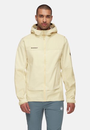 Man draagt een lichtbeige capuchonjas met "Mammut"-logo en grijze broek met een wit cirkelvormig logo op de linker dij.