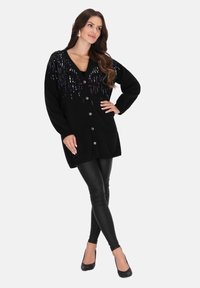 Schwarze Strickjacke mit tiefem V-Ausschnitt, verziert mit glänzenden lila Pailletten, mit Knopfverschluss und übergroßen Ärmeln, kombiniert mit schwarzen Leggings.