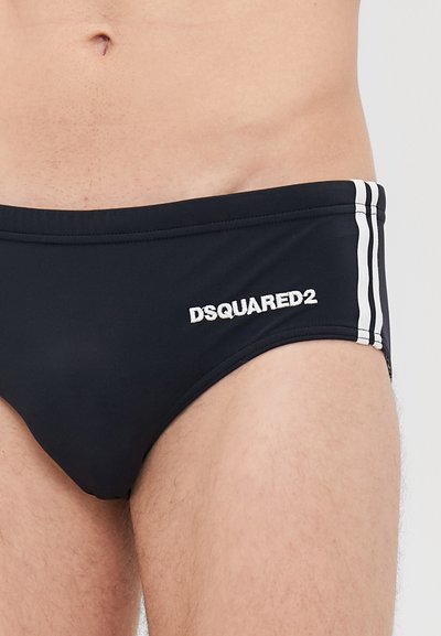 Männlicher Oberkörper in schwarzen Schwimm-Badehosen mit weißem DSQUARED2-Logo und zwei weißen Streifen an der Seite.