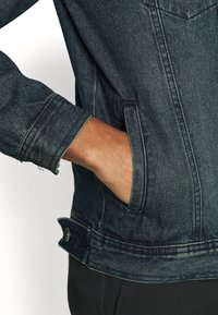 Dunkelblaue Jeansjacke mit einer Vordertasche, Knopfakzenten und sichtbaren Nähdetails. Das Design zeigt eine lässige Passform und einen strukturierten Stoff.