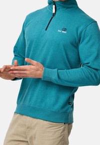 Sudadera de forro polar turquesa con un cuello alto, cierre de media cremallera, puños de canalé y un pequeño logo bordado en el lado izquierdo del pecho.