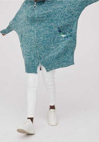 Turkusowy, dziany, oversize'owy kardigan z zamkiem, z wzorzystymi kieszeniami. Nosi się go na białych, dopasowanych spodniach i białych trampkach.