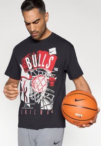 Czarny bawełniany t-shirt z dużym czerwono-białym napisem „BULLS” i wzorem kosza do koszykówki. Model trzyma pomarańczową piłkę do koszykówki Nike.