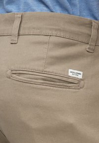 Béžové nohavice s vreckom na zadnej strane a horizontálnym otvorom. Viditeľná nálepka s nápisom "JACK & JONES EST. 1990." Hladká textúra látky.