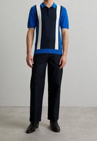 Modrý a tmavě modrý polo tričko s bílými vertikálními pruhy, krátkými rukávy a žebrovaným límcem, doplněné černými širokými kalhotami a černými botami.