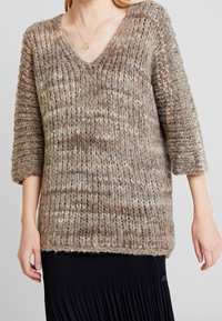 Brun och beige chunky stickad V-ringad tröja i ull med trekvartsärmar, bärs över en svart plisserad kjol.