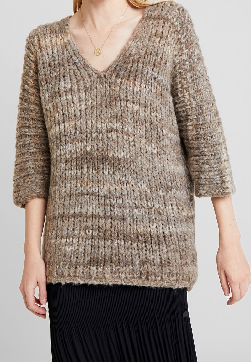 Brun och beige chunky stickad V-ringad tröja i ull med trekvartsärmar, bärs över en svart plisserad kjol.