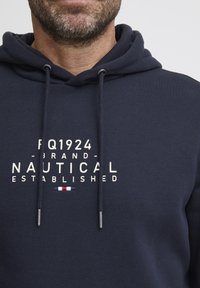 Sötétkék kapucnis pulóver, pamutból készült, elöl fehér nyomtatással "FQ1924 BRAND NAUTICAL ESTABLISHED" felirattal, valamint diszkrét piros, fehér és kék akcentussal.
