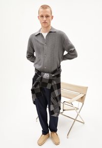 Cardigan en tricot gris avec col, superposé à une chemise ; pantalon bleu marine, chaussures beiges et écharpe à carreaux noirs et gris nouée à la taille.