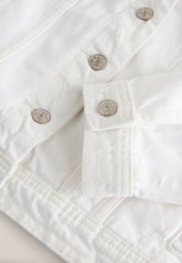 Biała kurtka dżinsowa zbliżenie z metalowymi guzikami, wzmocnionym przeszyciem i fakturowanym wykończeniem. Posiada detal w postaci podwiniętych mankietów.