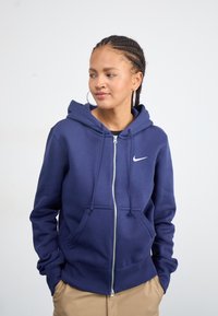 Tummansininen vetoketjullinen huppari, joka on valmistettu pehmeästä materiaalista. Siinä on naruhuppu, kengurupussit ja valkoinen Nike-logo rintakehässä.