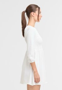Witte jurk met lange mouwen, textuur op het lijfje, gerimpelde taille en uitlopende rok. Eenvoudig ontwerp en lichtgewicht materiaal.