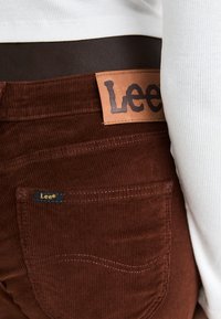 Bruine corduroy broek met een leren label waarop "Lee" in zwarte letters staat. Zichtbare stiksels en een klein logo op de zak.