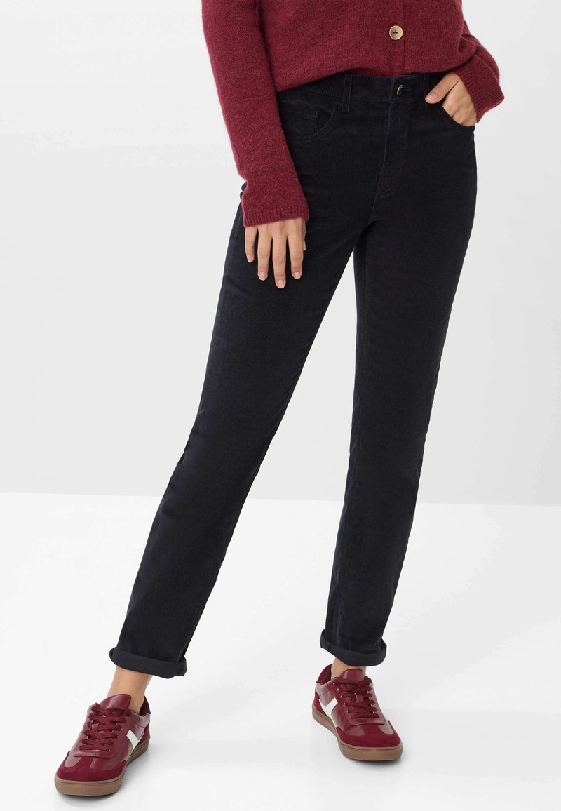 Schwarze Jeans im Straight-Leg-Schnitt, aus texturiertem Material. Kombiniert mit burgunderfarbenen Sneakers und einem kastanienbraunen Pullover mit Knopf.