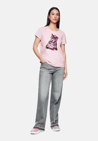 Frau in einem pinken T-Shirt mit einem schwarz-pinken Katzenmotiv, grauen weit geschnittenen Jeans und pink-weißen Turnschuhen, die mit einer Hand in der Tasche posiert.