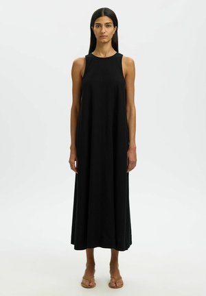 Vrouw staand, draagt een mouwloze zwarte jurk tot op de enkels en beige open-toe sandalen tegen een effen lichte achtergrond.