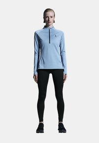 Himmelblaues Langarmshirt mit einem Viertelreißverschluss-Design, kombiniert mit schwarzen Leggings. Verfügt über eine figurbetonte Passform und ein Logo auf der Brust.