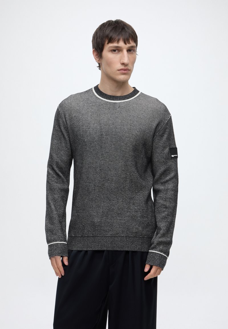 Jeune homme aux cheveux courts et foncés, portant un pull en maille gris avec des bordures blanches et un pantalon noir ample sur un fond clair uni.