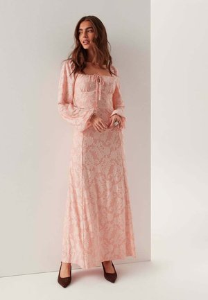 Kvinde iført en lang, lyserødblomstret blondekjole med ballonærmer og firkantet udskæring, stående mod en simpel hvid væg, iført mørke spidse sko.