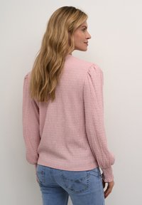 Maglietta lunga a maniche con texture rosa chiaro e maniche arricciate, abbinata a jeans blu aderenti. La vista posteriore evidenzia il tessuto soffice e i dettagli discreti.