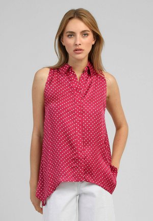 Femme portant un chemisier rouge sans manches à pois blancs et un pantalon blanc, debout devant un fond gris uni.