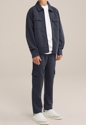 Marineblaue strukturierte Jacke mit zwei Vordertaschen, kombiniert mit passenden Hosen mit Cargotaschen und weißen Turnschuhen.