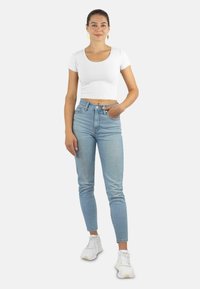 Weißes, kurzes T-Shirt, hellblaue, hochtaillierte Jeans und weiße Sneakers. Das Model steht mit einer Hand in der Jeans-Tasche vor einem neutralen Hintergrund.