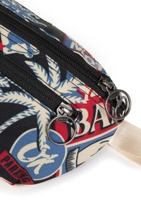 Lærredstaske med et multicolor print af palmeblade og logoer. To sorte lynlåse med runde metaltræk og en beige rem.