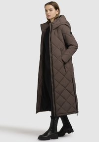 Brun, quiltet lang frakke med hætte, sideskaber og sorte accentdetaljer. Lavet af polstret materiale og har en tætsiddende silhuet.