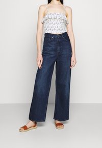 Žena oblečená v bílé květované crop top s volánky, tmavomodrých širokých kalhotách s vysokým pasem a hnědých sandálech s otevřenou špičkou, stojící na jednobarevném pozadí.