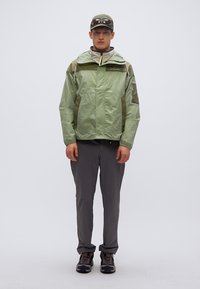 Ανοιχτοπράσινο αδιάβροχο μπουφάν με φερμουάρ, δύο πλαϊνές τσέπες και λεπτομέρειες από δίχτυ, συνδυασμένο με γκρι παντελόνι και σκούρα παπούτσια πεζοπορίας.