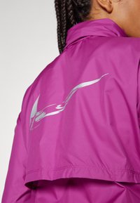 Femme aux cheveux tressés portant une veste violet vif avec un dessin d'oiseau argenté réfléchissant sur la partie supérieure du dos.