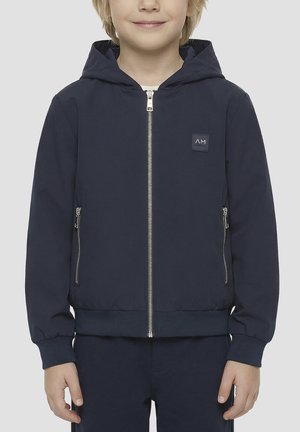 Niño con chaqueta azul marino con capucha, cremallera frontal y bolsillos laterales con cremallera, de pie frente a un fondo liso.