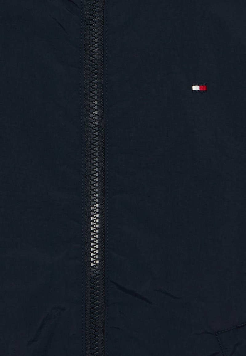 Veste marine avec une fermeture éclair centrale dotée d'une tirette texturée et un petit logo brodé en rouge, blanc et bleu sur la poitrine.