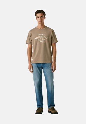 Giovane uomo in piedi che indossa una t-shirt beige con grafica "Notting Hill", jeans blu e scarpe casual marroni su sfondo bianco.