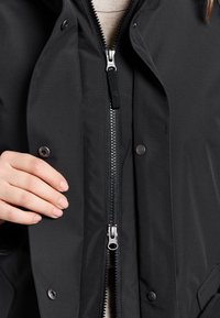 Svart ytterjacka med genomskinlig dragkedja, tryckknappar och textilstruktur. Har en hand på dragkedjan för detalj.