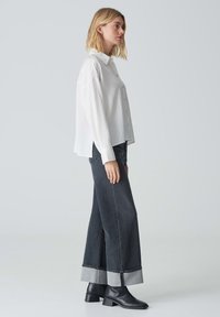Weißes, gestreiftes Button-Up-Hemd mit Kragen, kombiniert mit weit geschnittenen, dunklen Jeans mit umgeschlagenem Saum. Schwarze Stiefeletten runden den Look ab.
