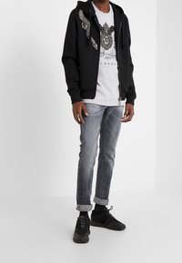 Sudadera negra con cremallera y diseño decorativo de alas plateadas en el hombro izquierdo, llevada sobre una camiseta gráfica gris y combinada con pantalones vaqueros gris claro y zapatillas negras.