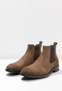 Botas Chelsea de ante marrón con paneles laterales elásticos, punteras redondeadas y suelas de goma negra. Cuentan con un tacón bajo para mayor estabilidad.